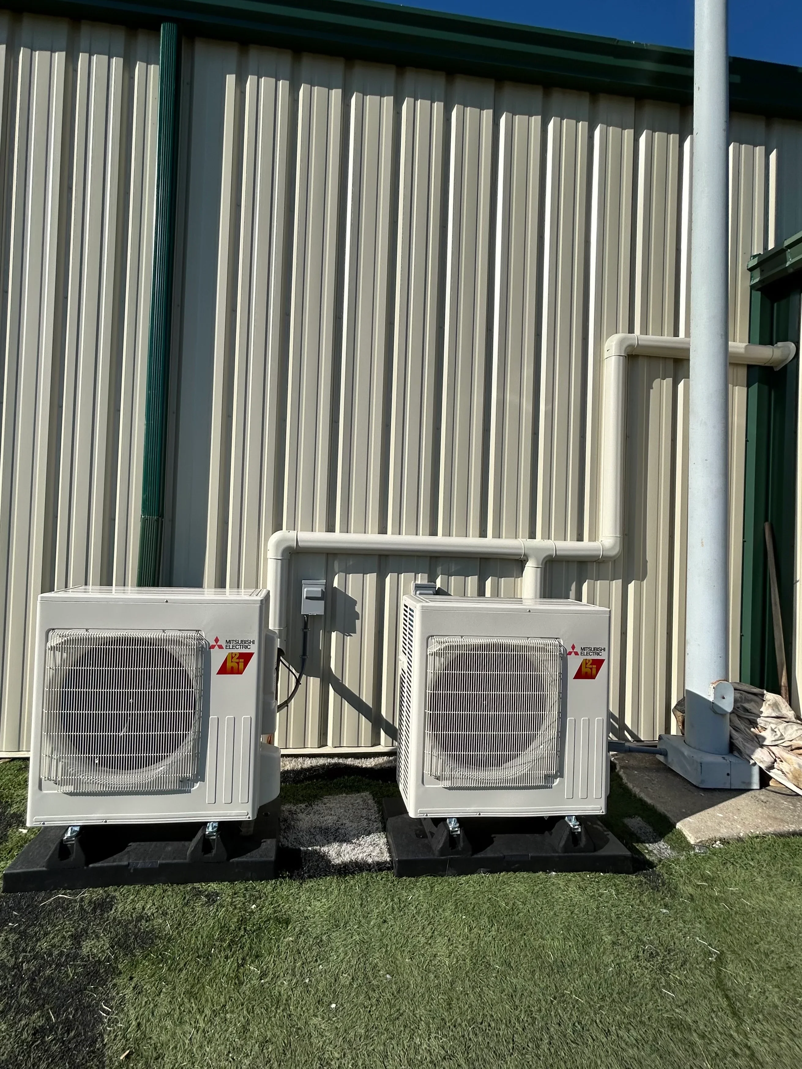 Two white Mitsubishi heat pump units on concrete pads with interconnected refrigerant piping and PVC drainage lines