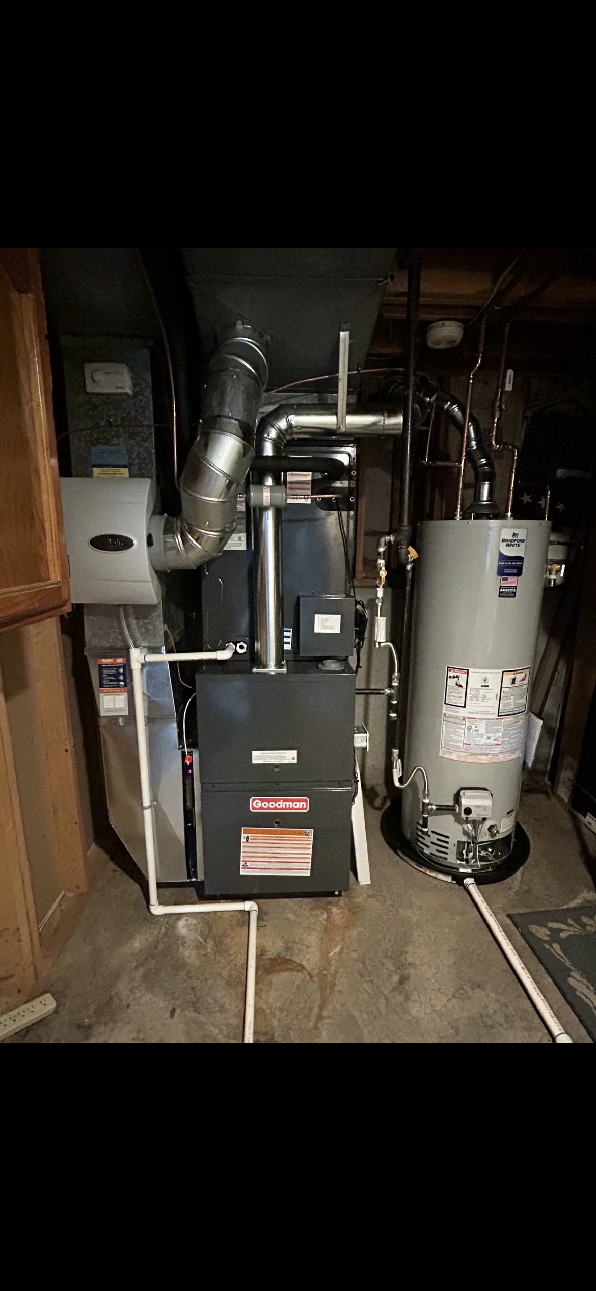 Goodman furnace with metal ductwork, gas water heater, and white PVC piping in basement installation
