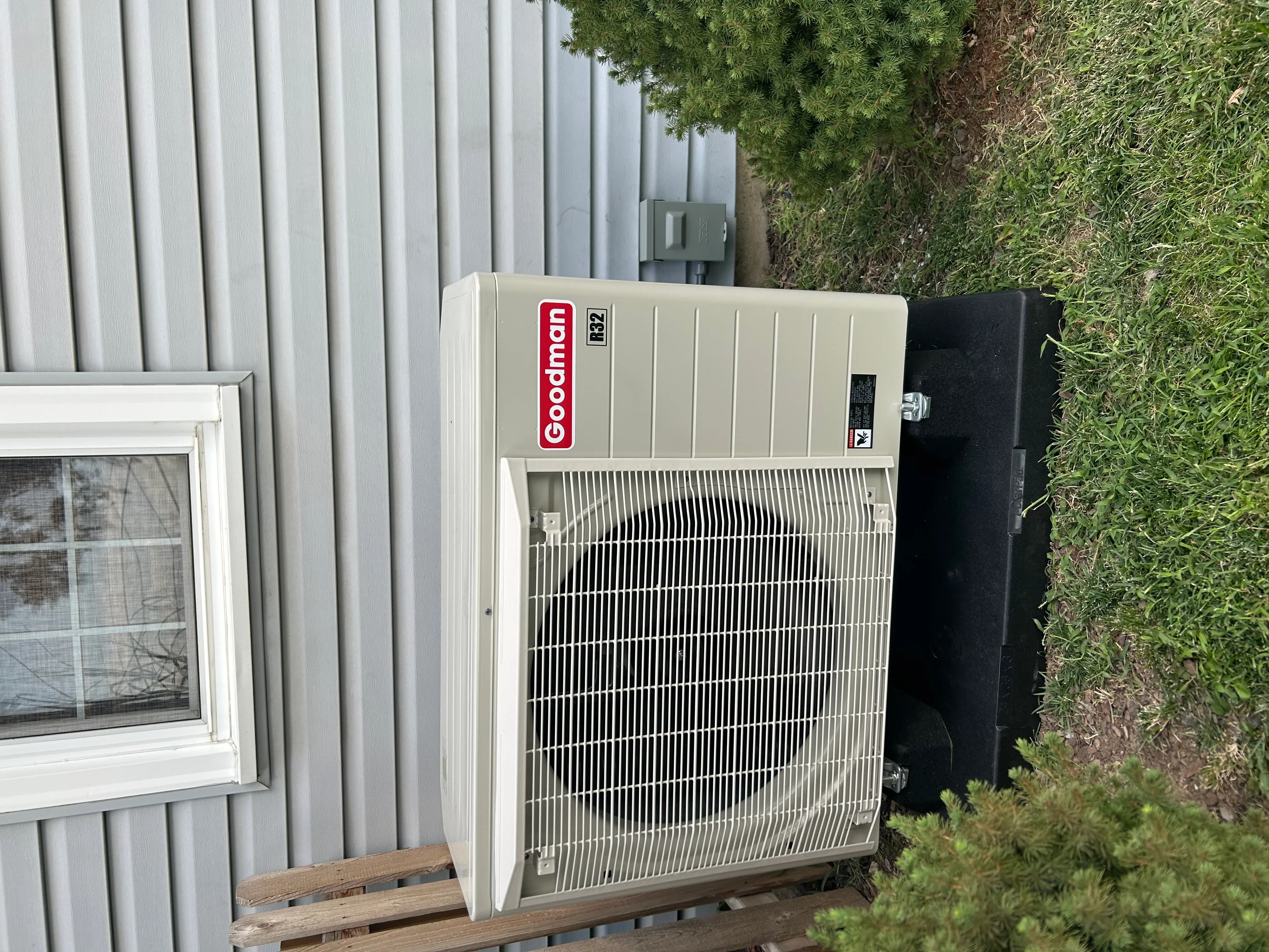 Goodman R22 heat pump unit installed on wooden pad with aluminum coil and metal siding backdrop